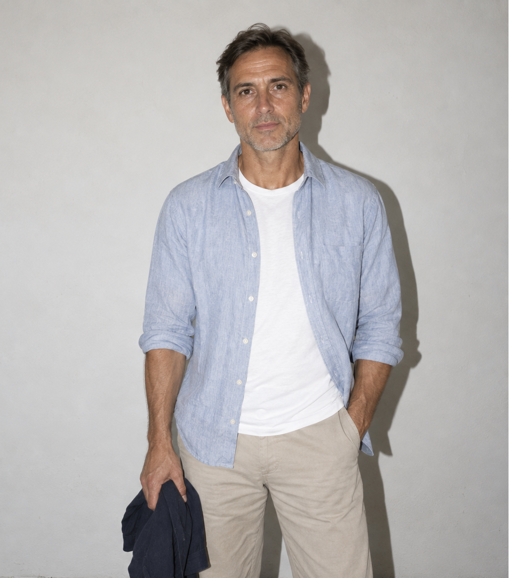 A man wearing a light blue linen button-down shirt over a white t-shirt, paired with beige trousers, standing against a plain light gray wall and holding a dark navy shirt in his hand.